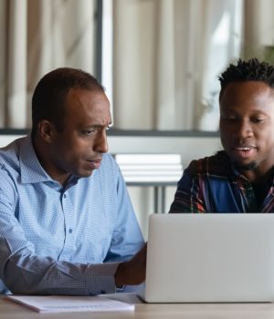 Two African American employees working on project together, using laptop pointing at screen, discussing strategy and sharing ideas at meeting, colleagues involved in teamwork, cooperation concept